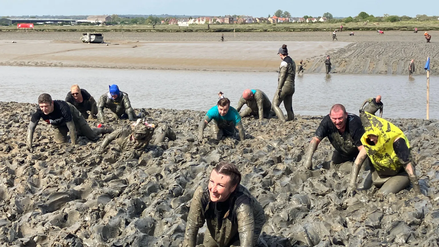 Men and women crawl through a muddy bank on a sunny day. They are covered in mud and some look exhausted as they make their way through. Other people can be seen behind them crawling through a flatter section of mud.