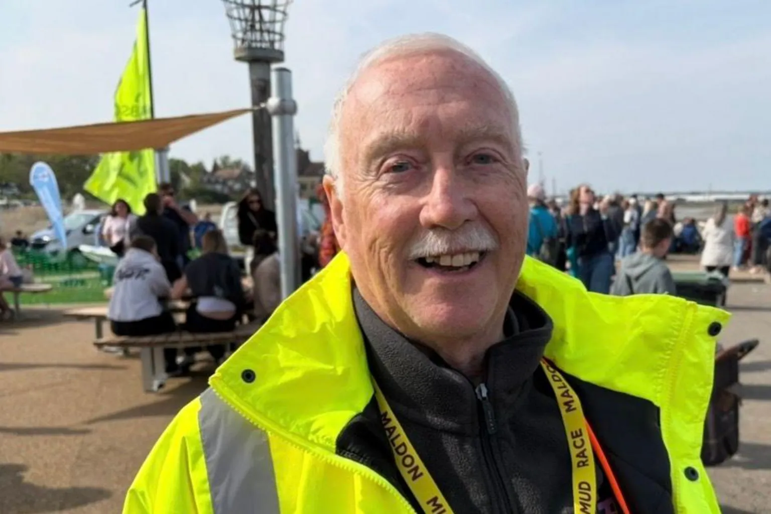 A man with grey hair and a grey moustache smiles at the camera. He wears a bright yellow hi-vis coat with a black jumper underneath and a yellow lanyard around his neck. People can be seen milling about behind at an event.