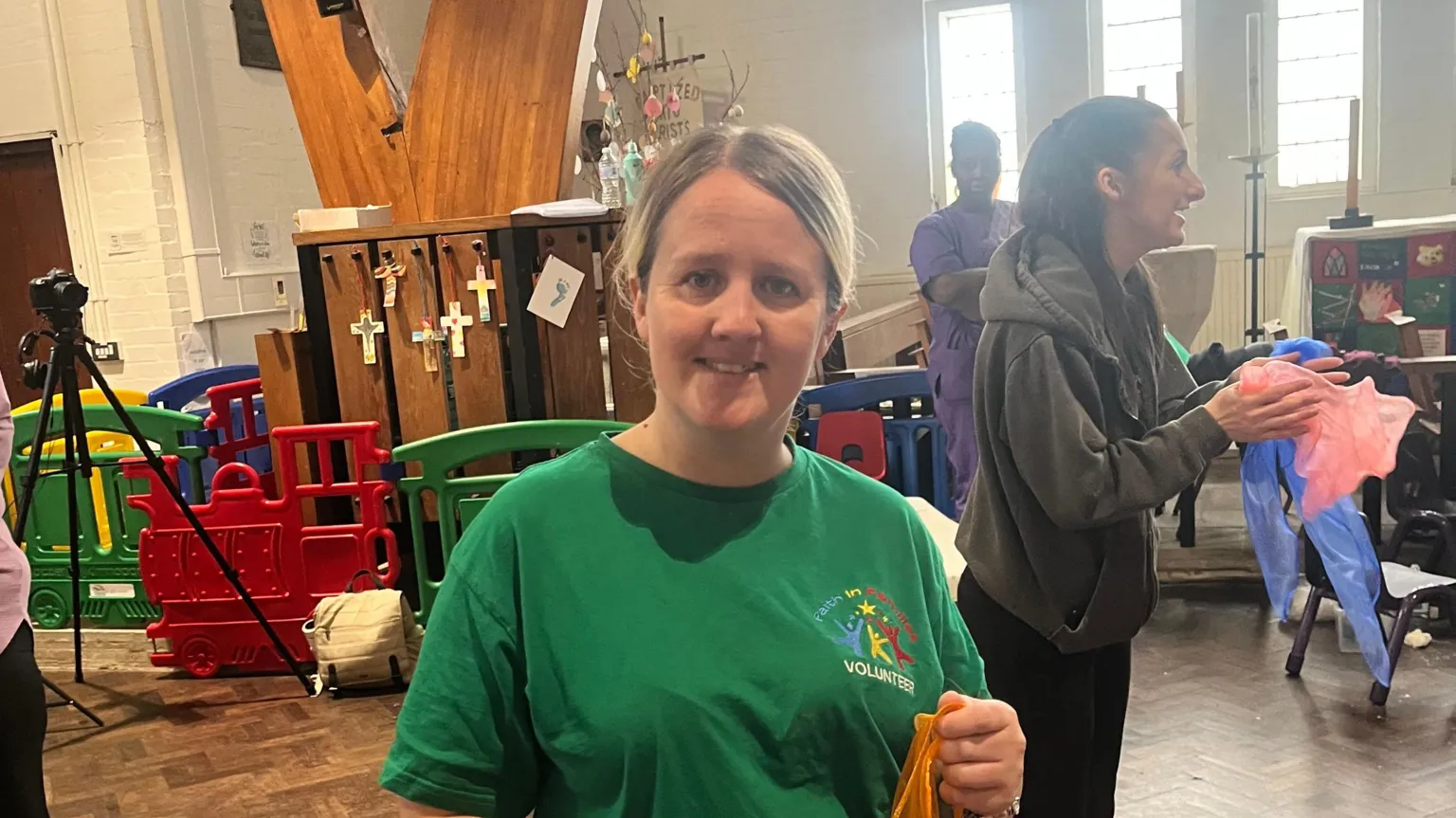 A blonde-haired woman in a green T-shirt stood in what looks like a church hall