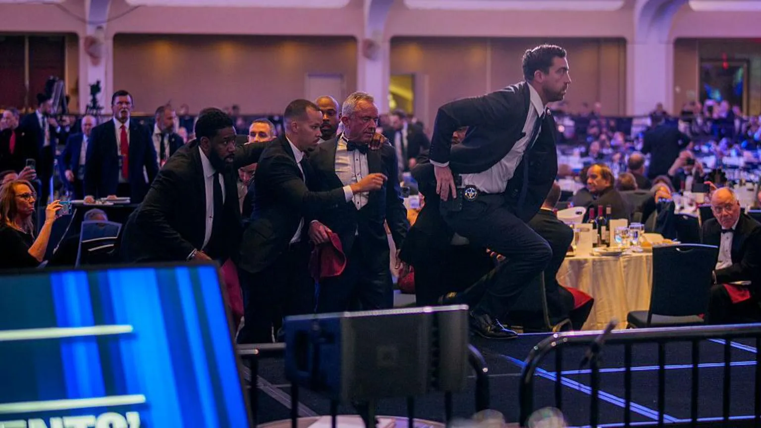  Kennedy surrounded by four men. One holds his shoulder and guides him while three run alongside, with one of them in front jumping onto a stage. They look frantic. People behind them either sit or stand, watching on 