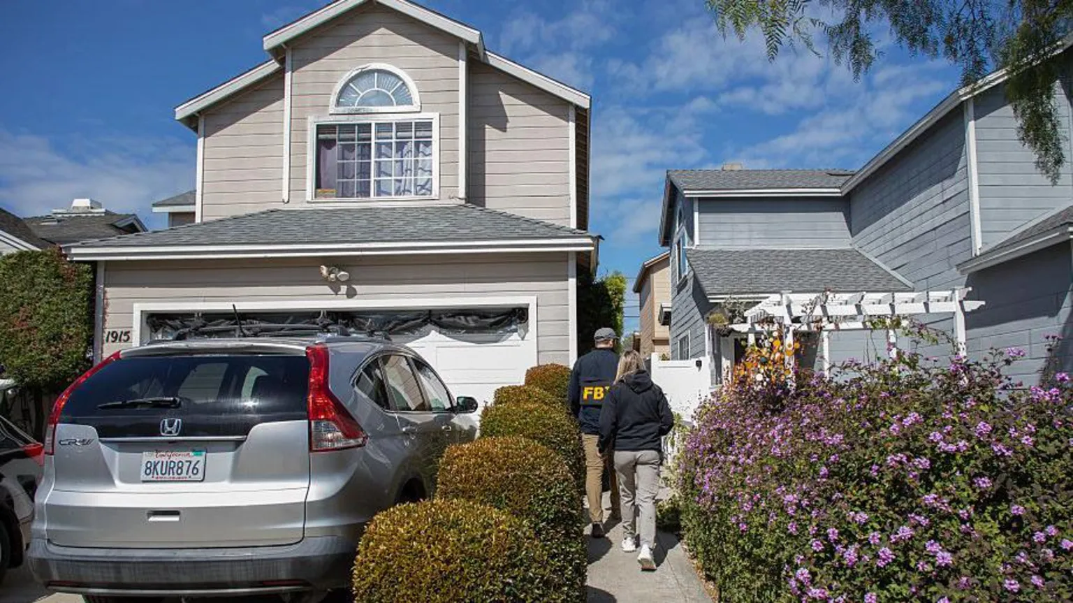  A man, wearing an FBI jacket, and a woman in a plain black jacket, walk towards a house with their backs to the camera. The house is two storey, light grey and close to the neighbour. The path is lined either side by manicured bushes, and a silver car is parked on the driveway. 