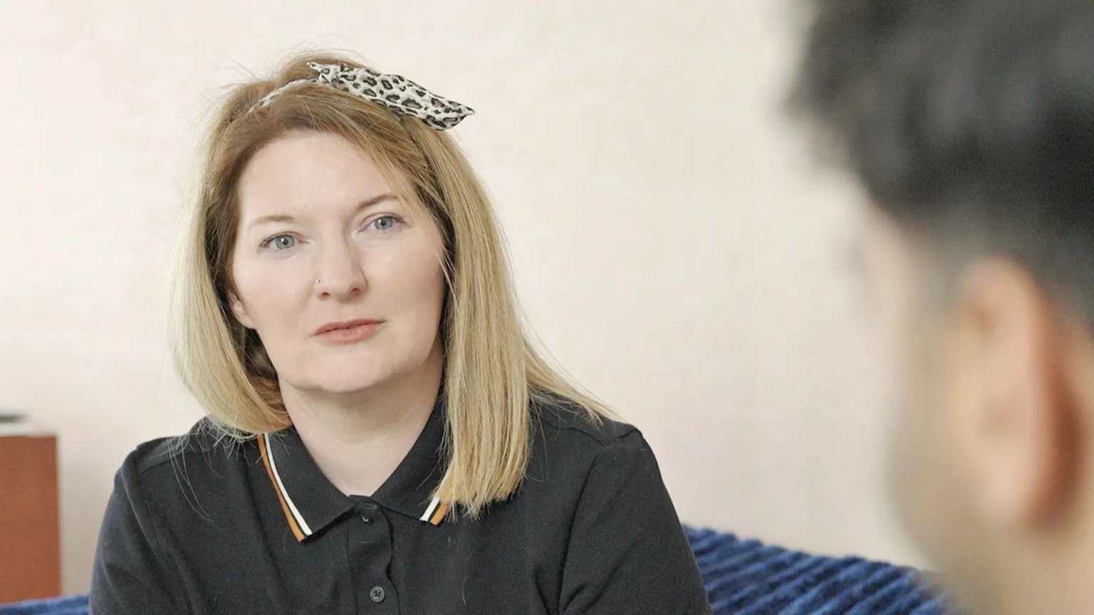 A woman looking towards the camera, wearing a black polo t-shirt, with straight fair hair and a leopard-print head band.