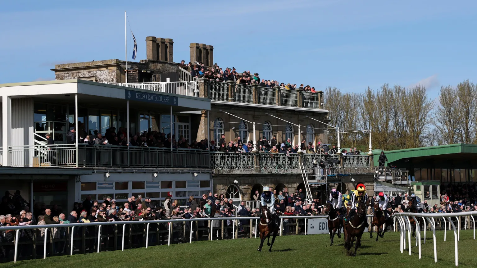  A packed grandstand at Kelso races with people also lined up behind the running rail watch as horses stream across the finish line