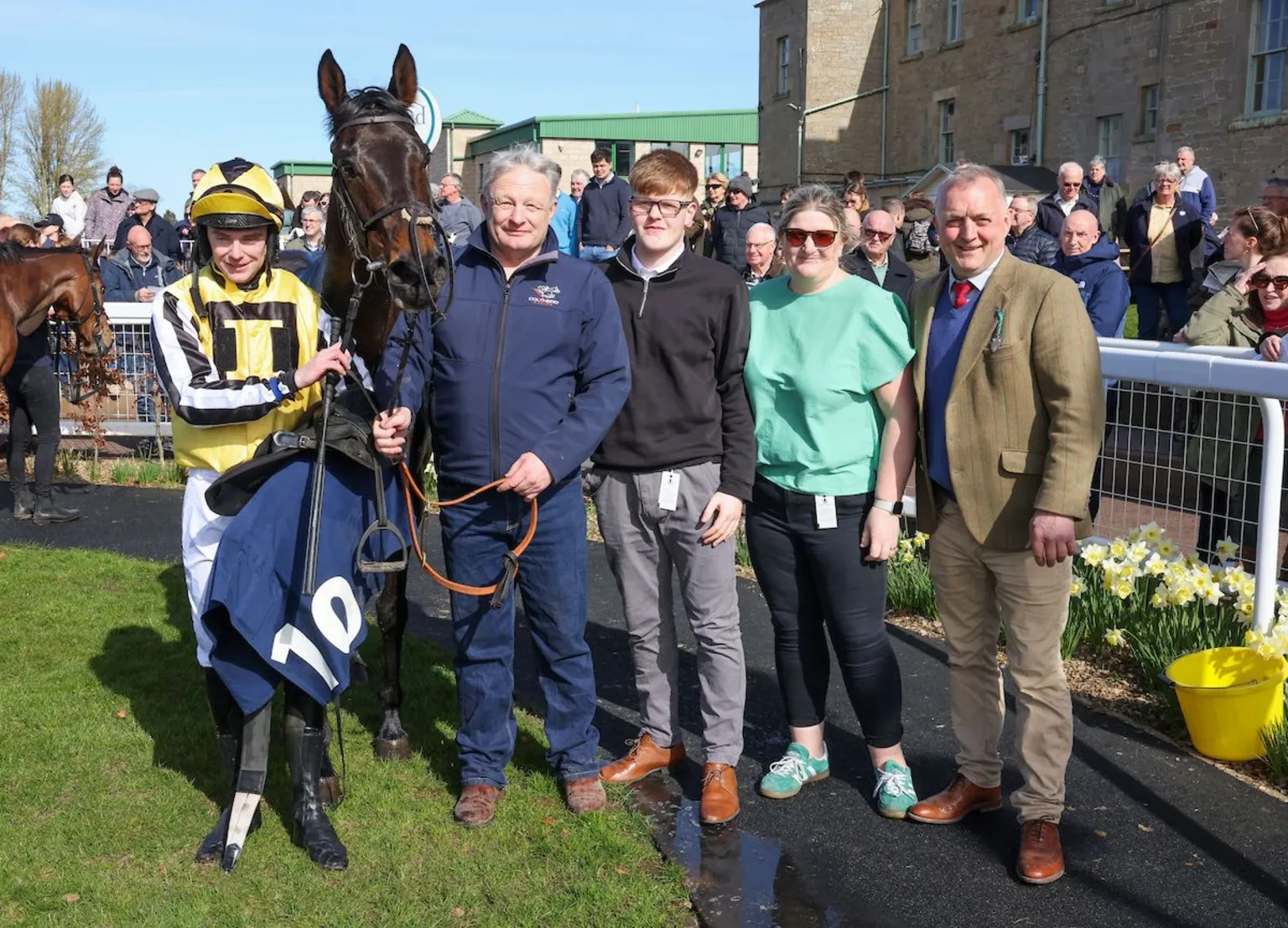 Grossick Photography A horse is held by the reins by a man with grey hair dressed all in blue and a jockey in yellow silks. A young man with ginger hair and a woman in a green top with black trousers and a man in a tweed jacket stand next to them looking into the camera.