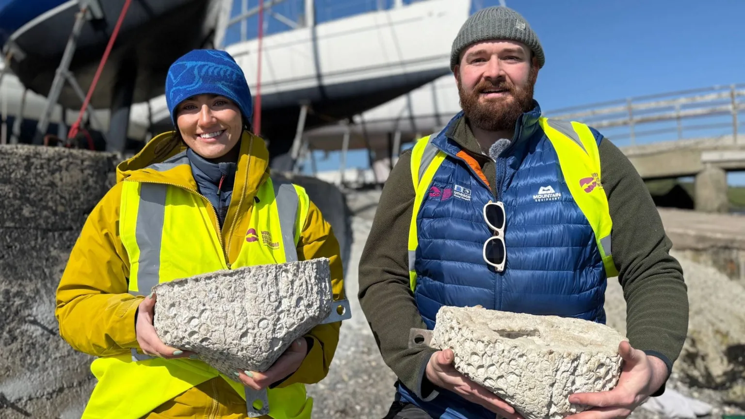Mourne Gullion and Strangford officers Anna and Grant holding grey rock concrete rock pools. Anna is wearing a blue hat, yellow mustard coat and a yellow high vis vest with Mourne Gullion and Strangford logo. Grant is beside her wearing a blue puffy gilet with sunglasses hanging on the zip. He is also wearing a high vis jacket but it is not fully covering him. He has a green jumper under his puffer. There are boats out of focus in the background and a large grey concrete wall. 