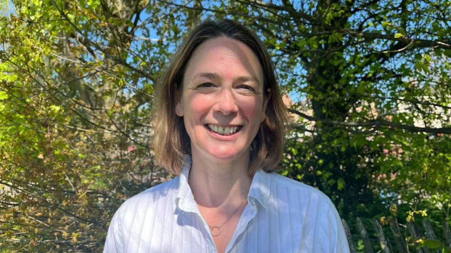 Ellen Knight/BBC Photo of Lynn looking into the camera and smiling. She has shoulder-length light brown hair, and is wearing a white collared shirt and a silver necklace with a circular pendant. She is photographed in front of green, leafy trees, with dappled sunlight falling on her face. The sky, seen beyond the trees, is bright blue and clear. 
