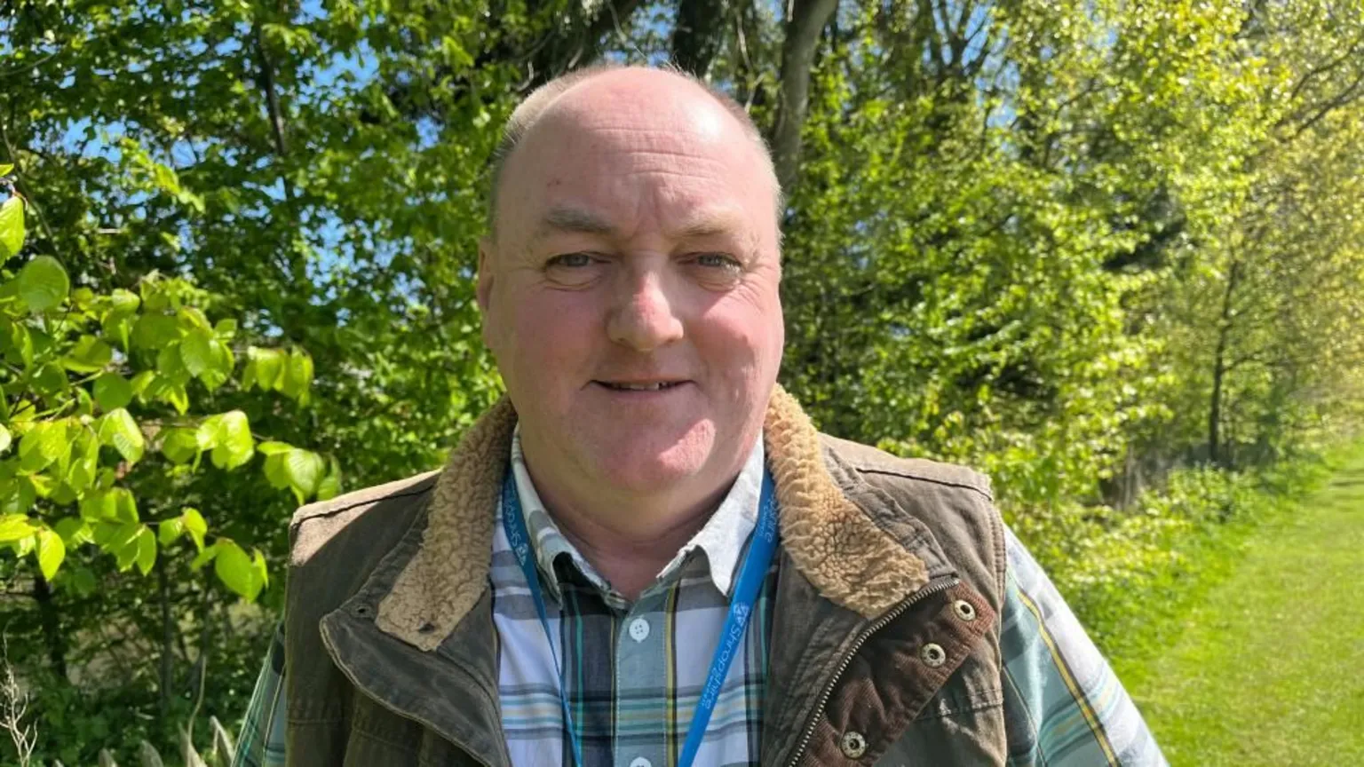 Ellen Knight/BBC Walker pictured looking into the camera and smiling slightly. He is wearing a green, yellow, and white tartan shirt with a brown waxed gilet over the top. He is also wearing a light blue Shropshire Council-branded lanyard. He is stood in front of a row of bright green trees and bushes. The sky is bright blue and there is strong sunshine. 