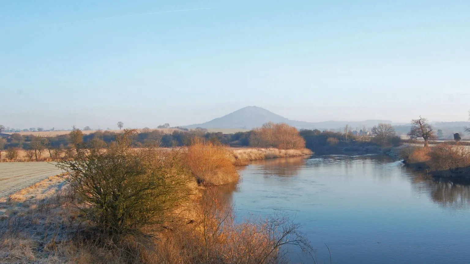  A wintery photograph of a river, with the Wrekin hill rising up in the distance, slightly obscured by cloud. The banks of the river are lined with trees and shrubbery, and the grass has a layer of frost on it. The sky is blue and slightly pink, suggesting the photo is taken near sunrise. 