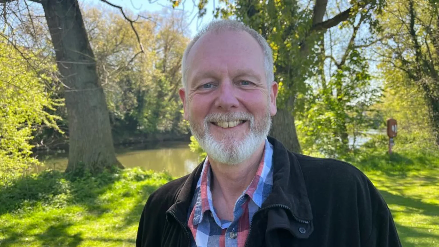 Ellen Knight/BBC Dave pictured looking into the camera and smiling. He is wearing a blue and red checked shirt, with a black fleece jacket over the top. He has short grey hair and a short grey beard. He is pictured stood on the banks of a river, with bright green grass and trees with green leaves lining the watercourse. The sky is bright blue, with strong sunshine casting dappled light on the ground. 