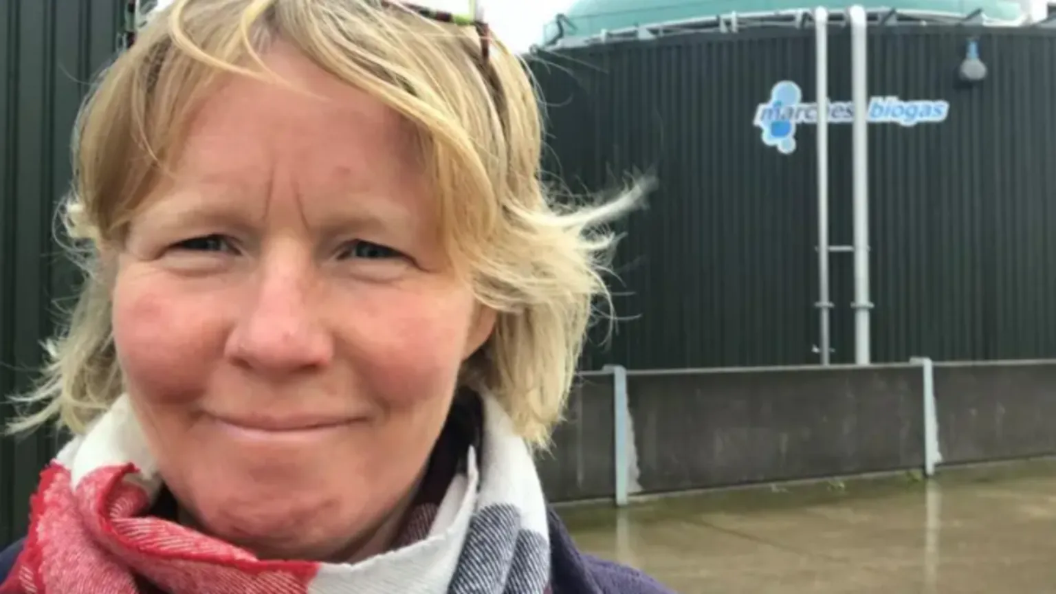 NFU Photograph of Kate taken in front of two huge biogas tanks on her farm. She has chin-length blonde hair, and is wearing a blue jacket and a red, white, and blue scarf. The tanks behind her have dark green corrugated iron containing them, with a concrete floor on the ground. 