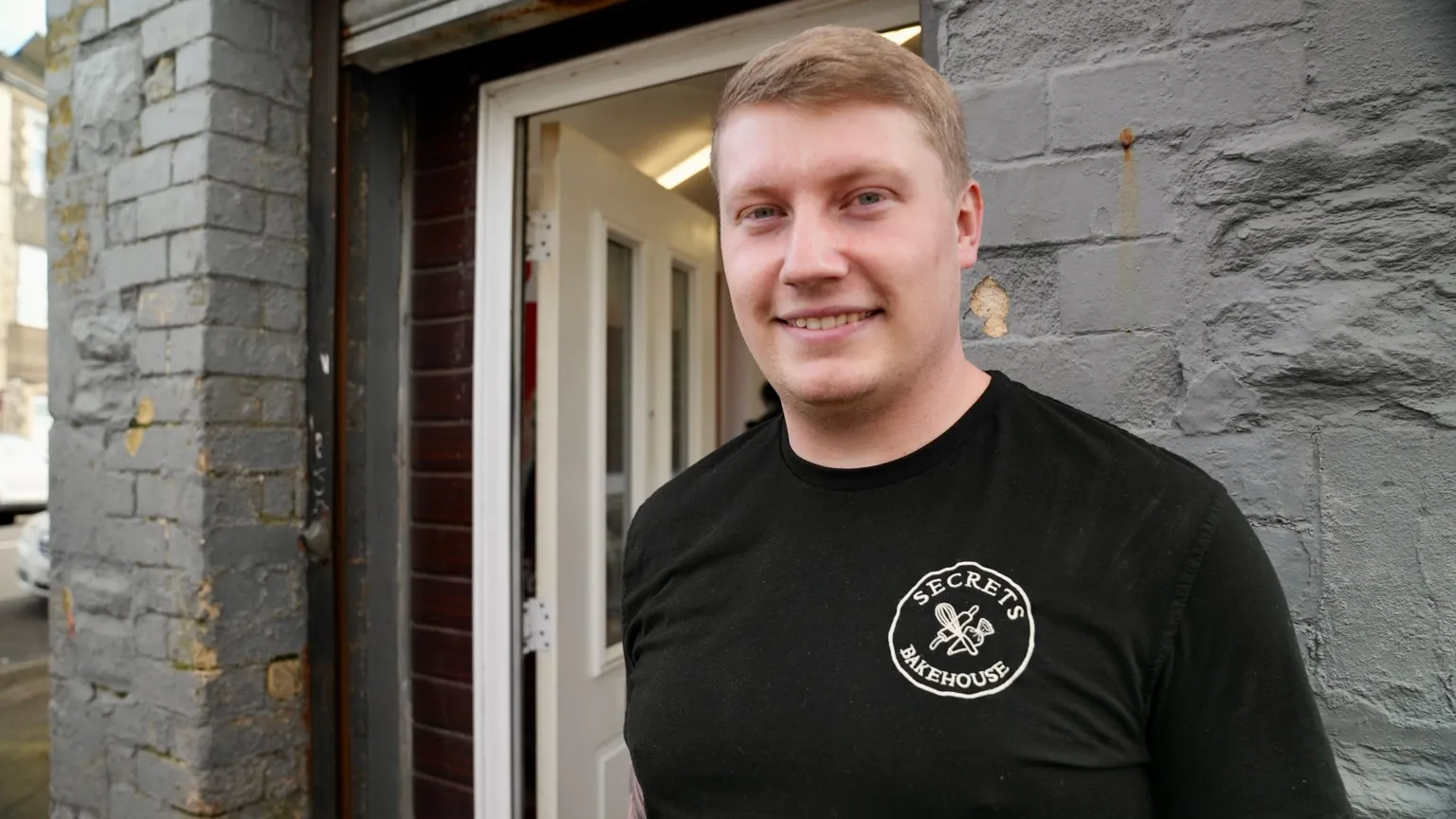 Kyle is wearing a black t-shirt, standing outside his bakery. He's looking at the camera and smiling.