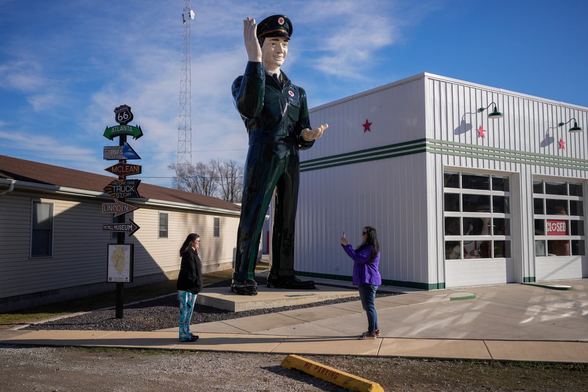 Two women take photographs by a giant sculpture of a gas station attendant in uniform 