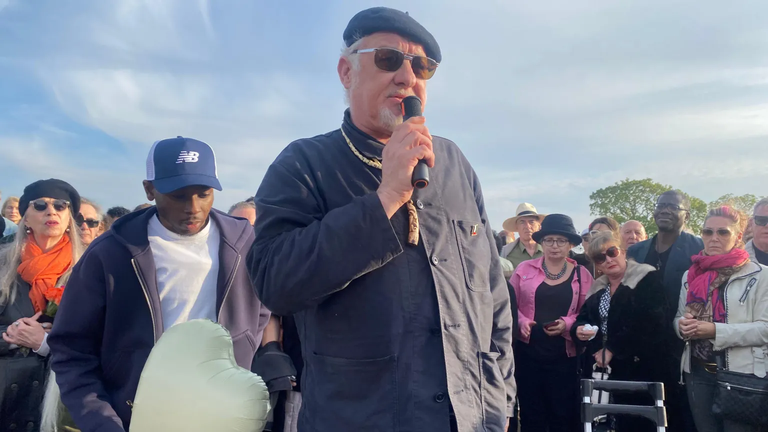 Chris Sullivan is shown speaking into a microphone outside surrounded by people. He is wearing a dark beret and sunglasses and dark shirt.