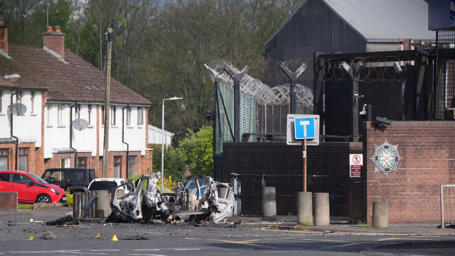Niall Carson/PA Wire The wreckage of a car bomb outside a police station, with a row of terraced houses and trees in the background. The car has been blown apart and lies on the road beside a set of pavement bollards. The station is protected by a large metal fence, topped with barbed wire, and a red brick wall. 