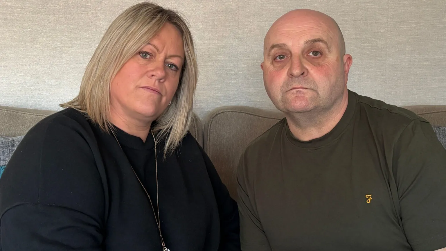 A woman with a short blonde bob wearing a black t-shirt and zip jacket. A ,am next to her is bald with facial stubble and wears a khaki green t-shirt with a small yellow logo on its breast.