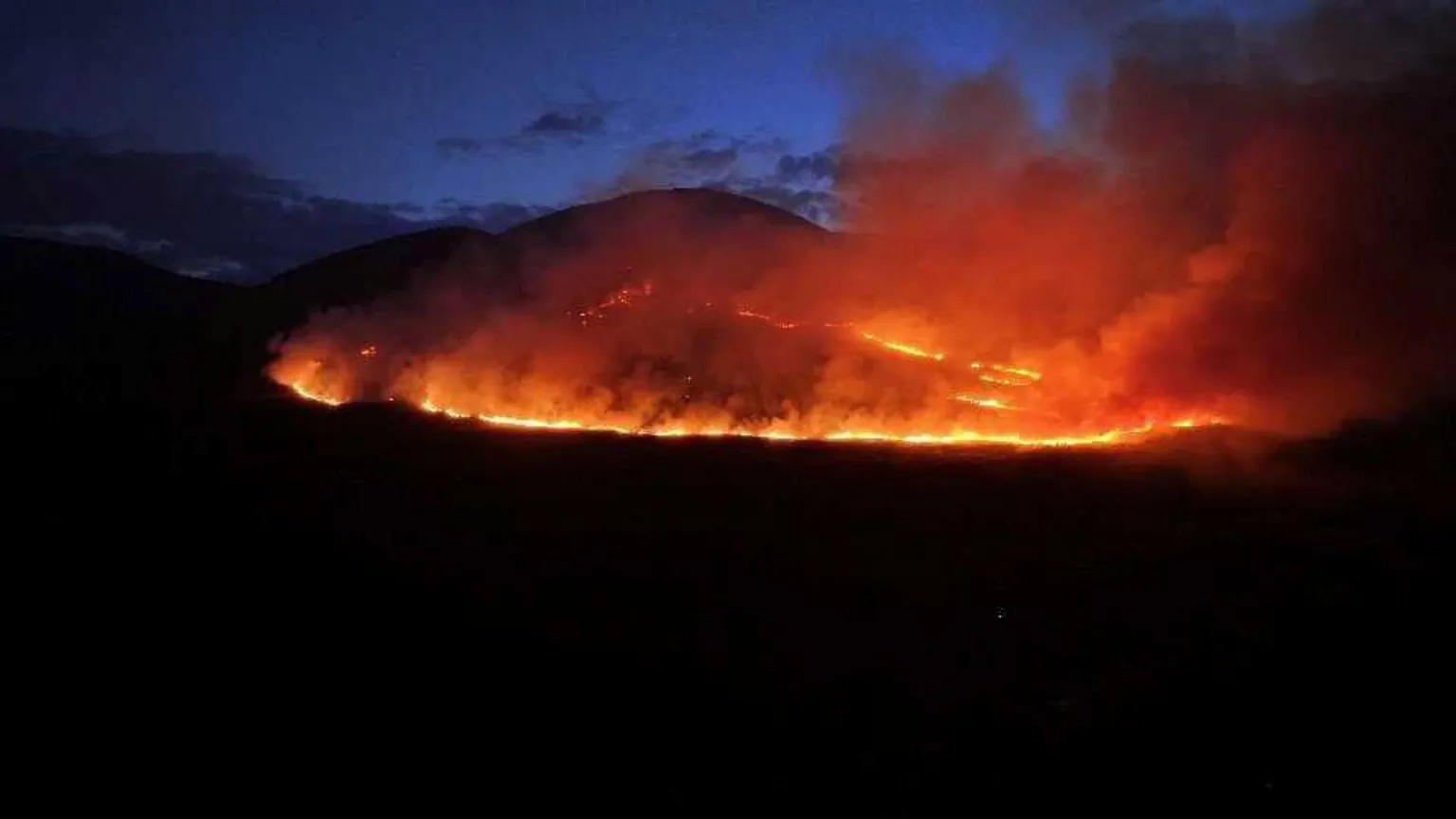 Jordan Dick A large wildfire on a mountain at night. Smoke is bellowing into the sky. The rest of the mountain is in darkness. The sky is dark navy with some clouds. 