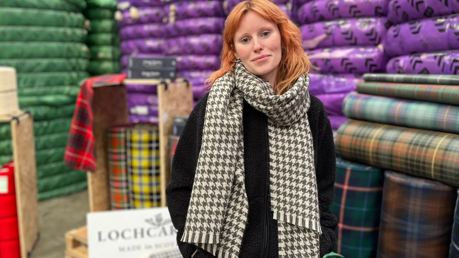 A girl with red hair, wearing a chequered scarf, stands in front of rolls of tartan