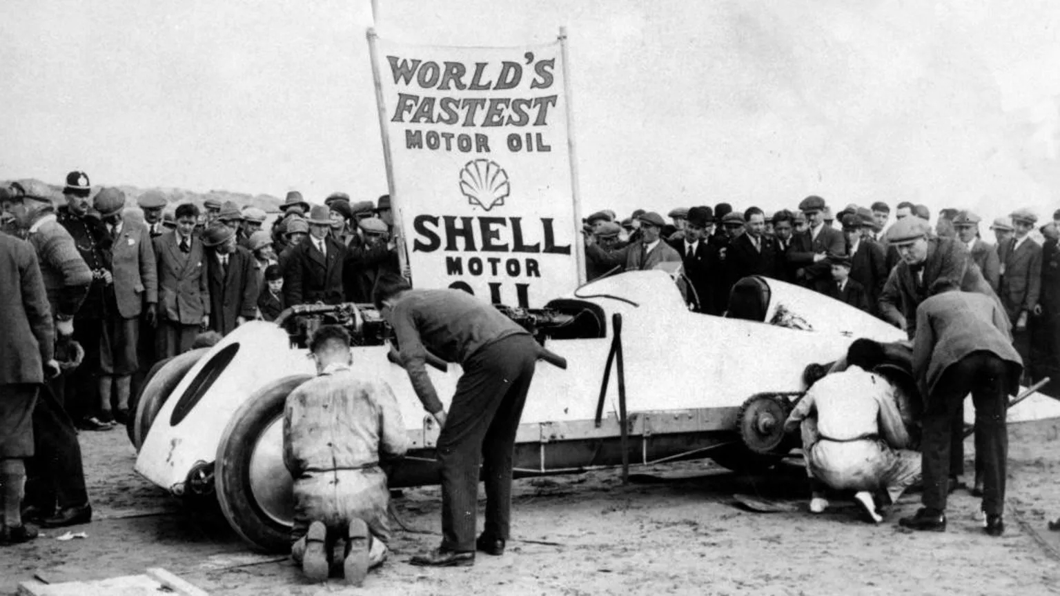  An archive picture of 'Babs' with mechanics at each wheel. There is a group of spectators behind the car watching. A sign behind the vehicle says 'Fastest motor oil Shell motor' with the Shell logo. 
