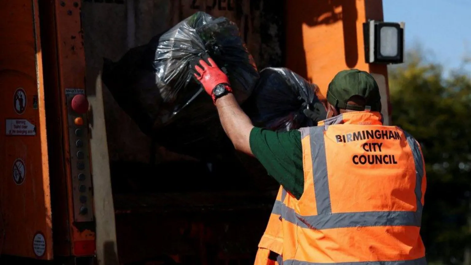  A man wearing a green hat, green shirt and an orange hi-vis jacket has his back to us as he throws two, full black bin bags into the back of a refuse vehicle.