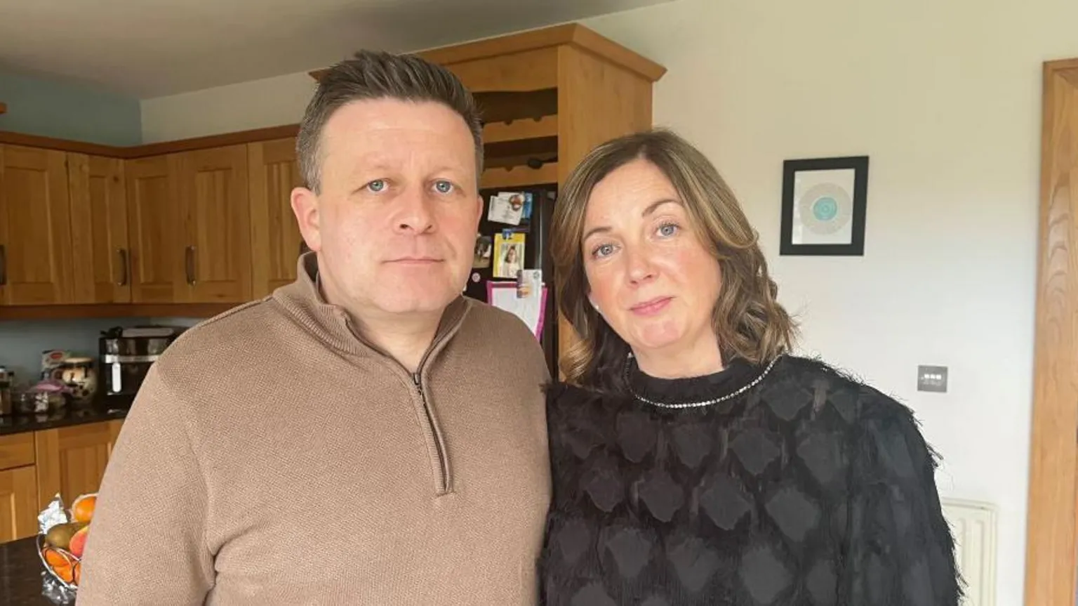 A couple standing in a kitchen looking into the camera. The man has a brown jumper on with a zip. He has brown hair and blue eyes. He is pictured beside his wife. She has brown hair with blonde highlights, blue eyes and is wearing a silver necklace. There are kitchen cupboards in the background. 
