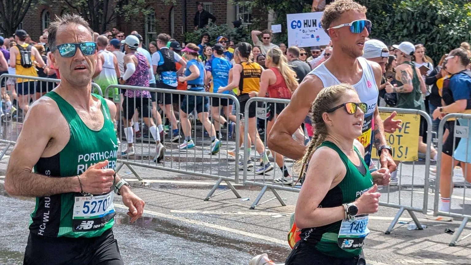 Gabriella Booth Two people running in the marathon wearing Porthcawl Runners branded tops. There is another runner just behind them wearing blue, reflective glasses. A group of runner are heading in the opposite direction in the background.