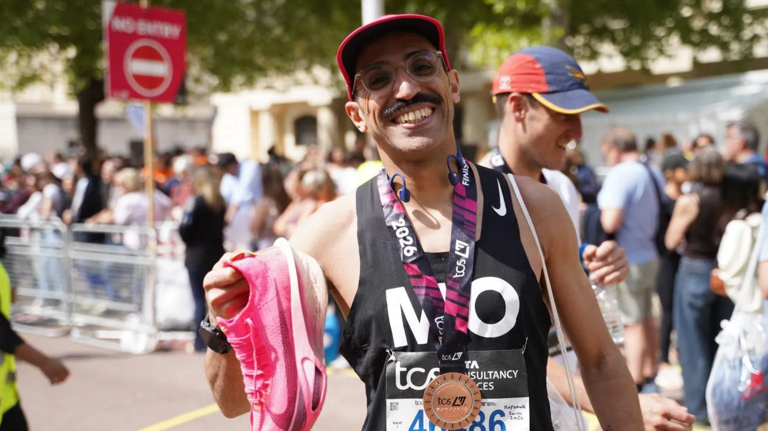 Mo Kachach A man smiles holding a pair of pink running shoes. He has a medal around his neck and and a hat protecting part of his face from the sun. There is another run stood behind him with a crowd of people watching the event on his left.