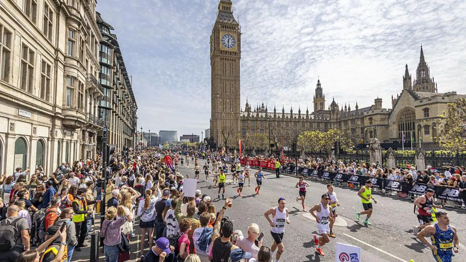  Runners can be seen with supporters on either side of the road behind barriers. Some hold signs, others have drinks in their hands. In the background you can see Big Ben attached to the Houses of Parliament.