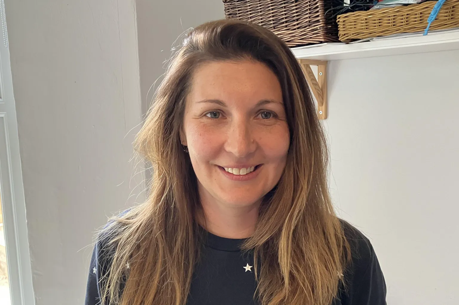 A woman of around 40 is smiling straight into the camera in a bright room with some shelving visible just over her head, with wicker baskets on. She is wearing a dark blue jumper with a pattern of while stars and has long light brown hair parted on the left and swept to the right.