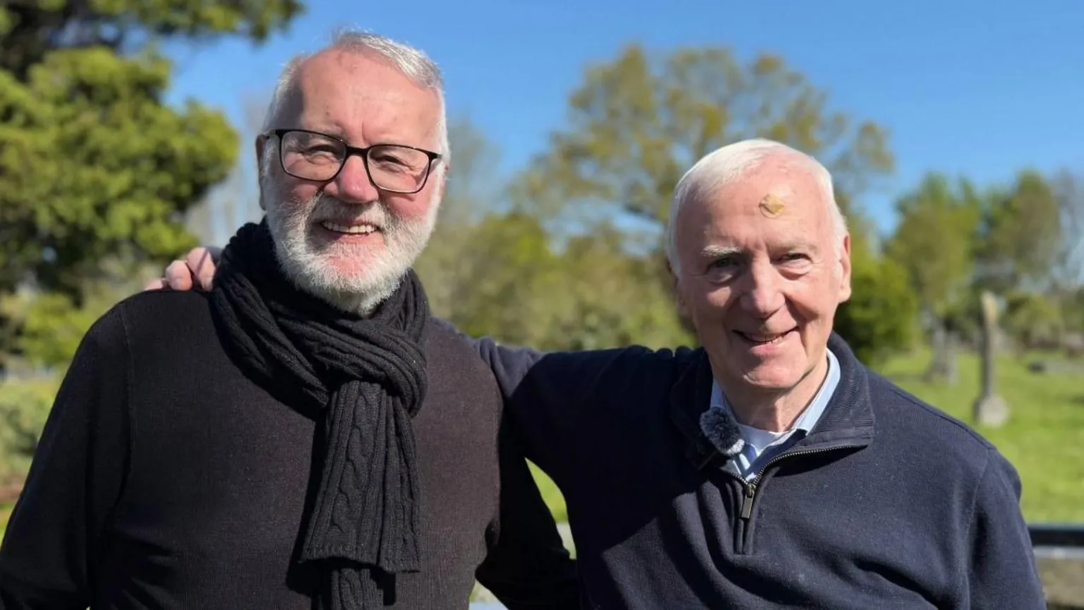 Two men with white hair. Man on the left hand side is wearing a black jumper with a black scarf. Man on right side is wearing a navy jumper. They are smiling at camera.