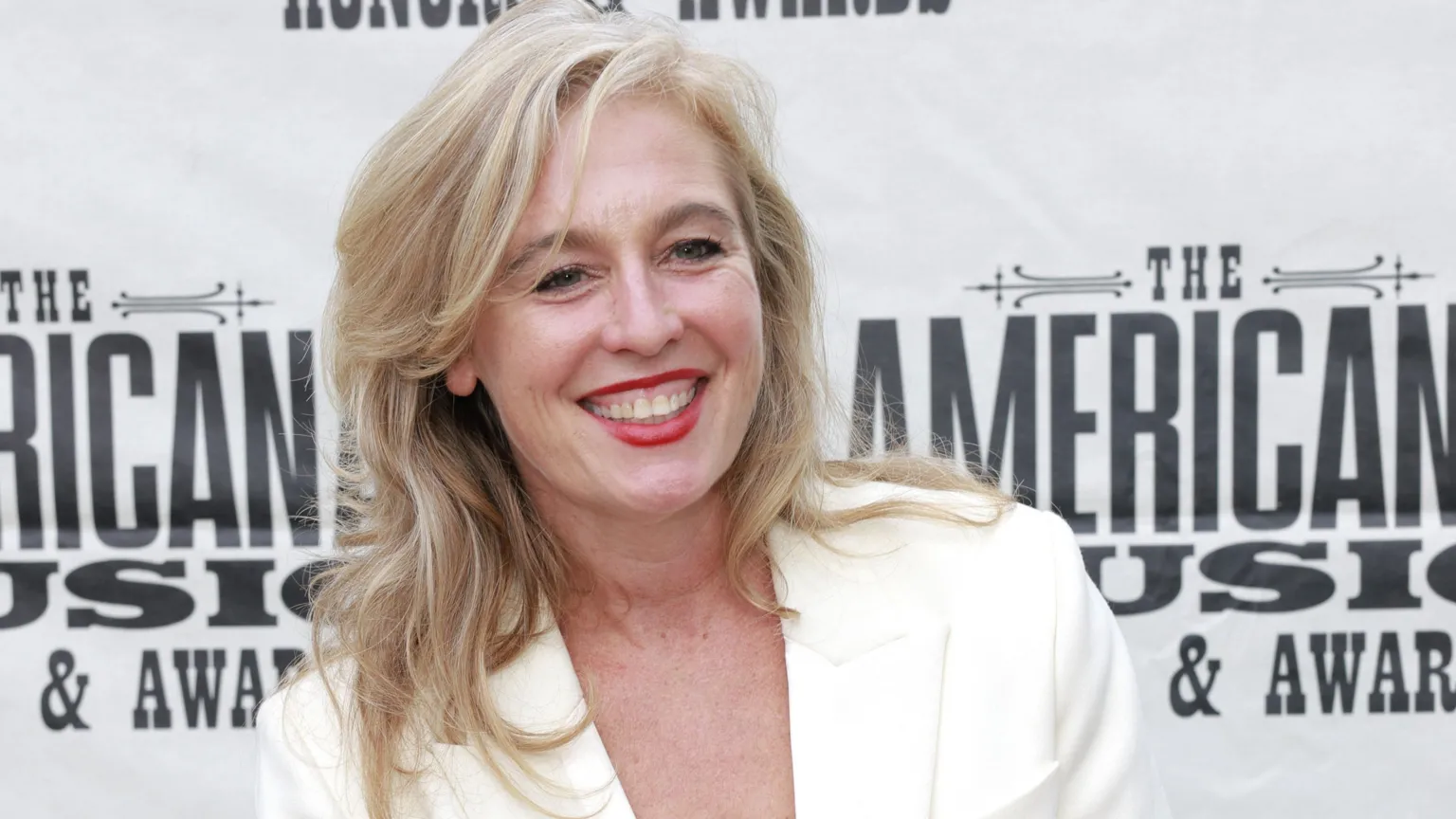  Tift Merritt on the red carpet of an awards ceremony in Nashville, Tennessee. Smiling, with blonde hair, she wears a white jacket.