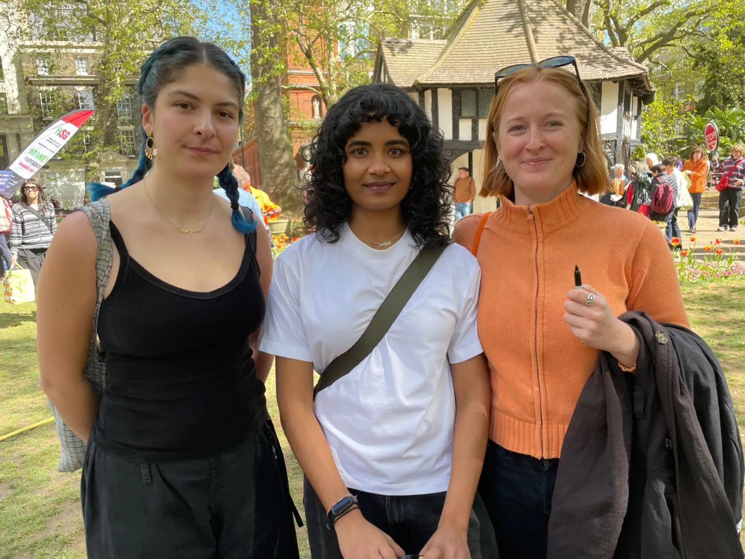 Three young women stand in a park, side by side. The woman on the left has blue highlights in her hair and wears a black vest top; the woman in the centre has dark shoulder-length curls and wears a white T-shirt; the woman on the right has shorter red hair and wears an orange zip-up jumper. 