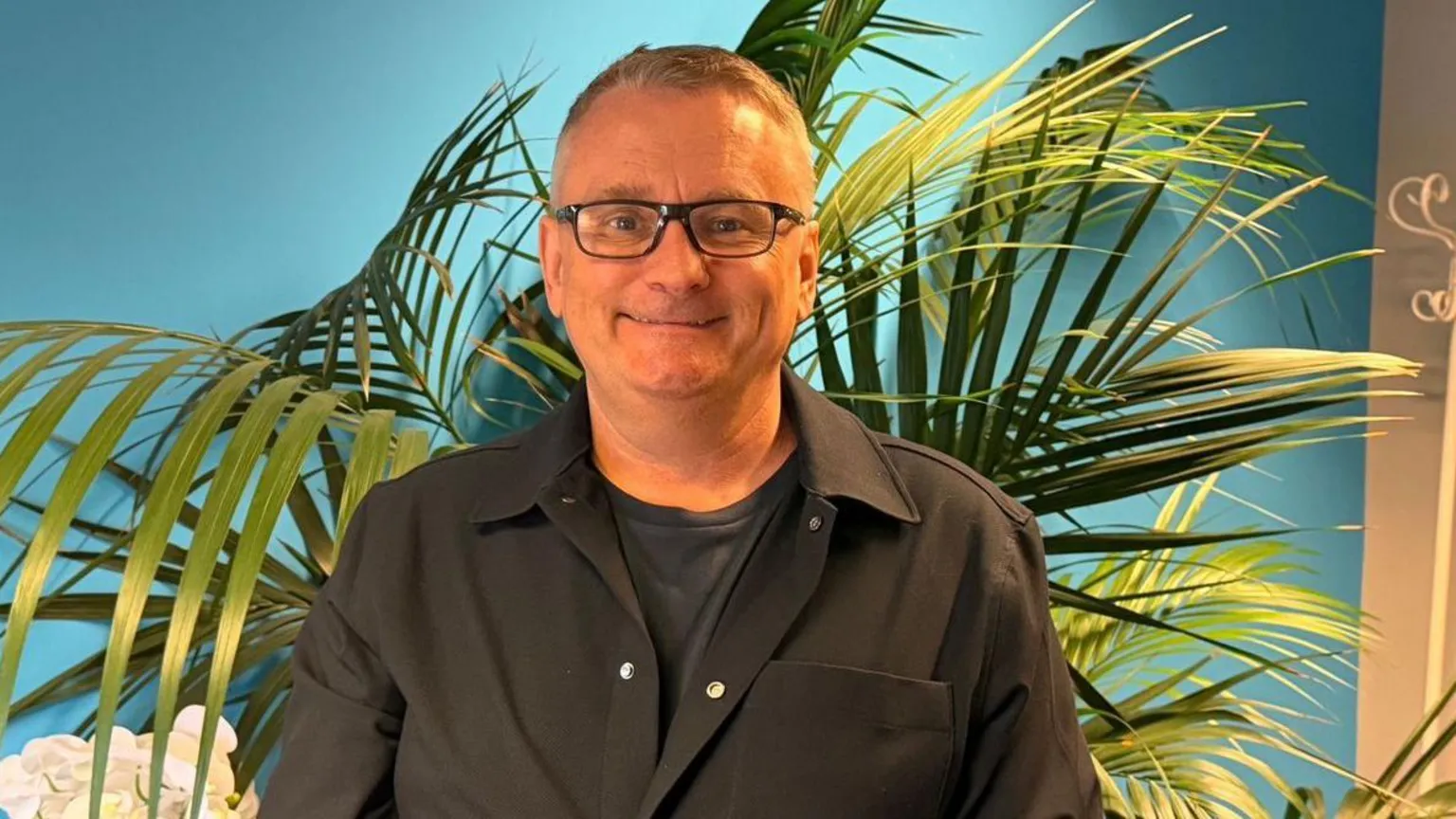 A middle-aged man in glasses stands against a blue background, with a large palm plant behind him. 