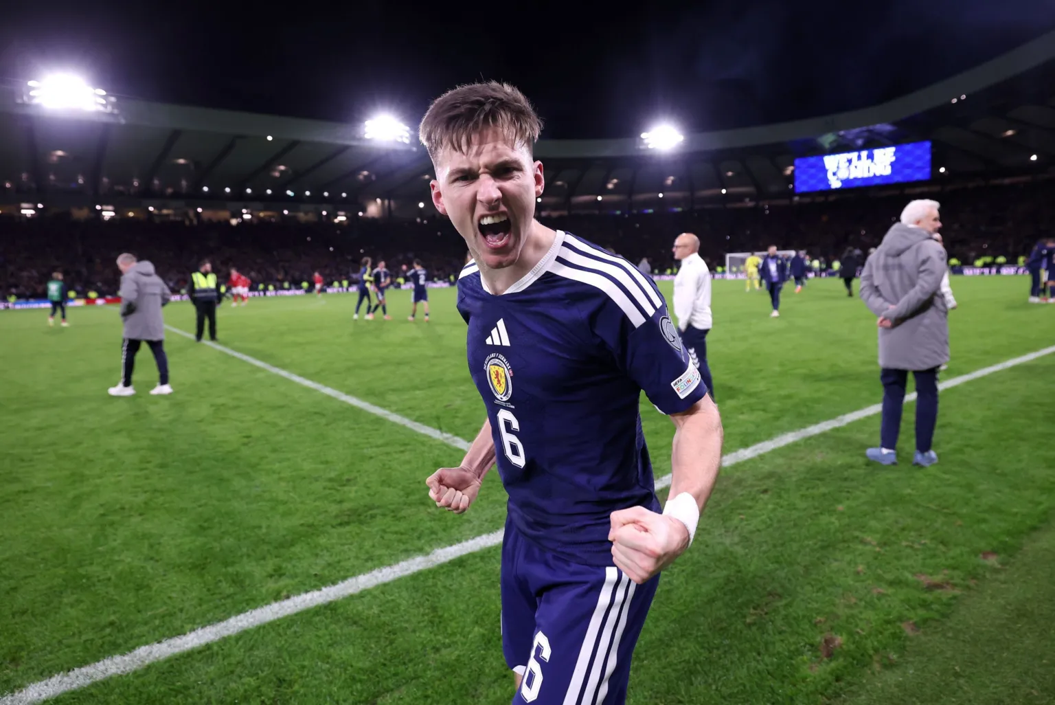  Kieran Tierney is wearing a blue Scotland kit and his mouth is open in a shout, his fists clinched as he celebrates at the side of the pitch.