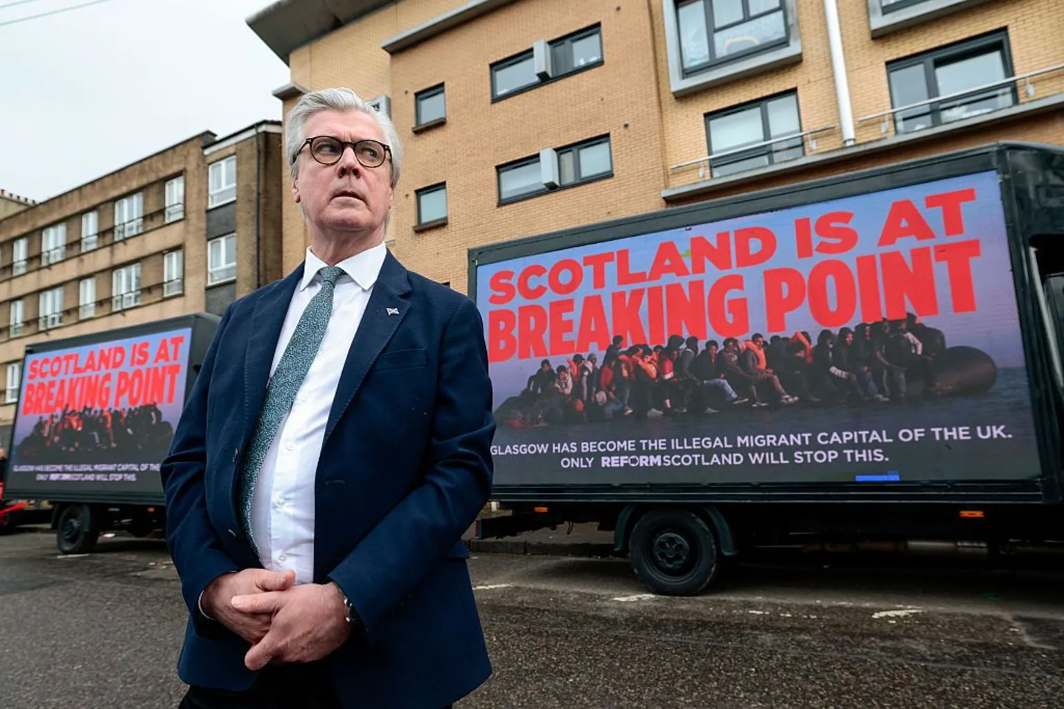  Reform’s Scottish leader Malcolm Offord wearing a blue suit, white shirt and green tie, attends a party election event where he stands in front of a van with the message 'Scotland is at breaking point' written on the side. 