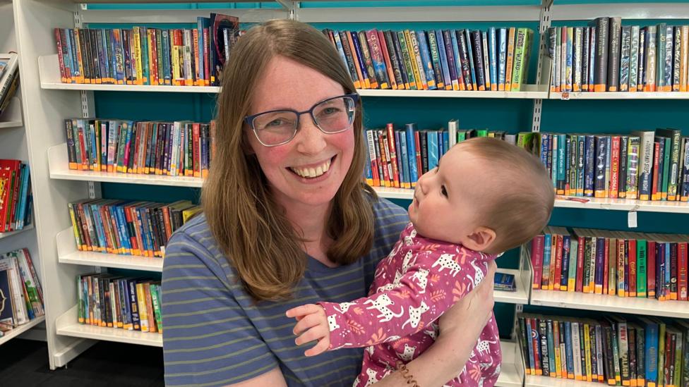 Mam ifanc mewn sbectol yn edrych tuag at y camera ac yn gwenu, mae ei phlentyn bach yn ei breichiau