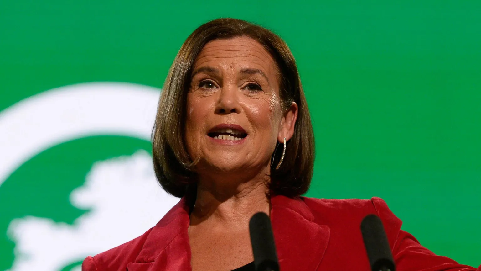  Mary Lou McDonald is pictured speaking at her party's annual conference. She has shoulder length brown hair and is dressed in a red blazer and back top, gesturing with her left hand. Behind her the backdrop is bright green and on the podium in front of her are the words 'Building Ireland's future'.