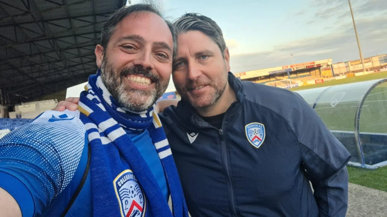 Andrea Lai Two men standing with their arms around each others shoulders, one is wearing a blue and white scarf the other a black jacket