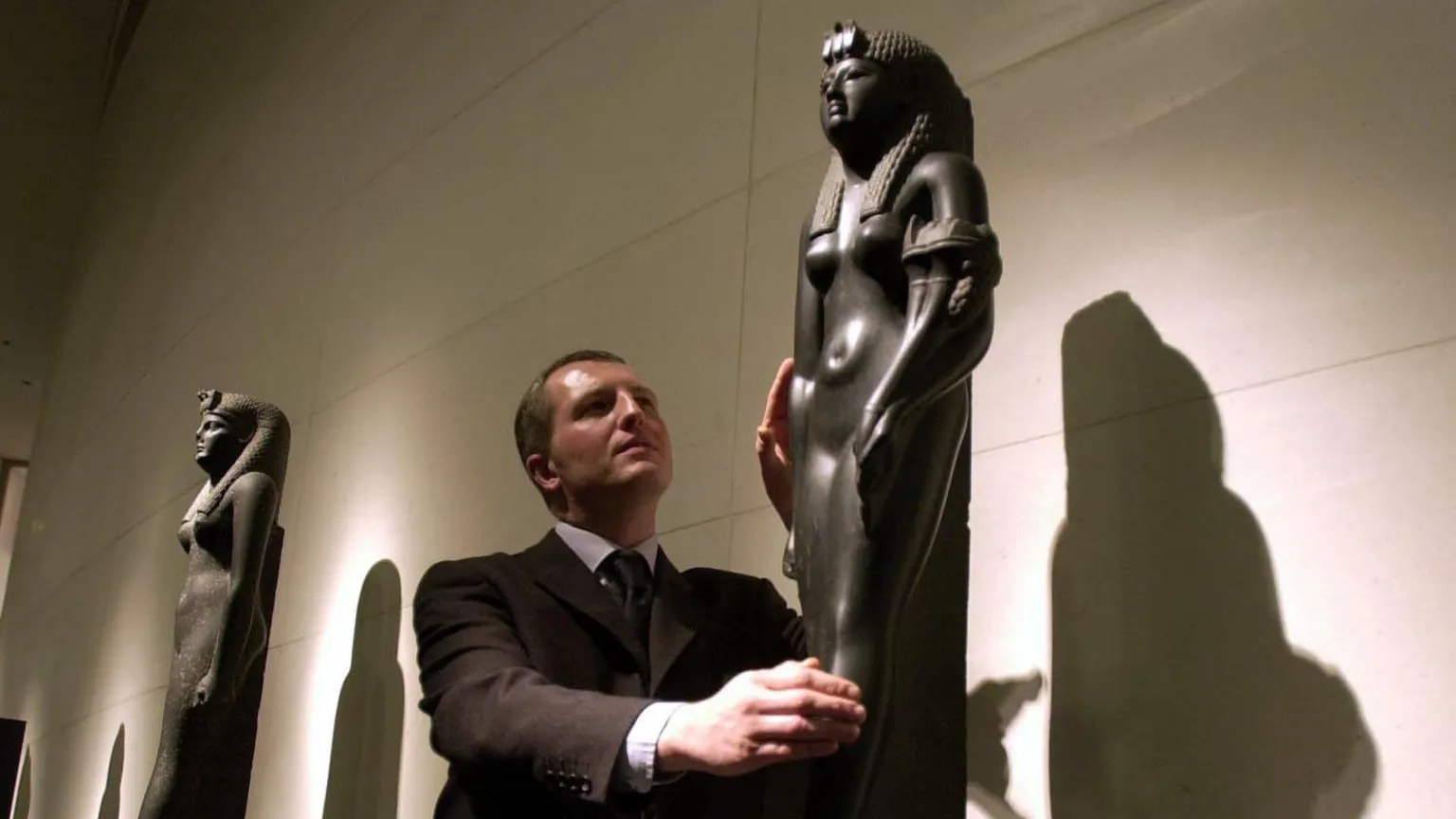 PA Dr Peter Higgs in brown suit in centre of image with a black basalt statue of Cleopatra VII, at the British Museum in 2001 Another statue of Cleopatra behind him.