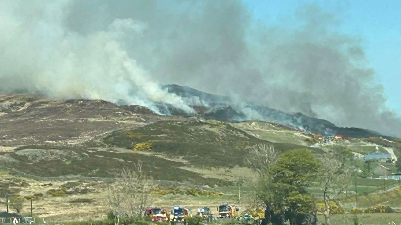 Shaun McAuley Smoke is rising from a mountain. Several fire engines are parked. Some trees and yellow bushes are growing. A few buildings are in the distance. 