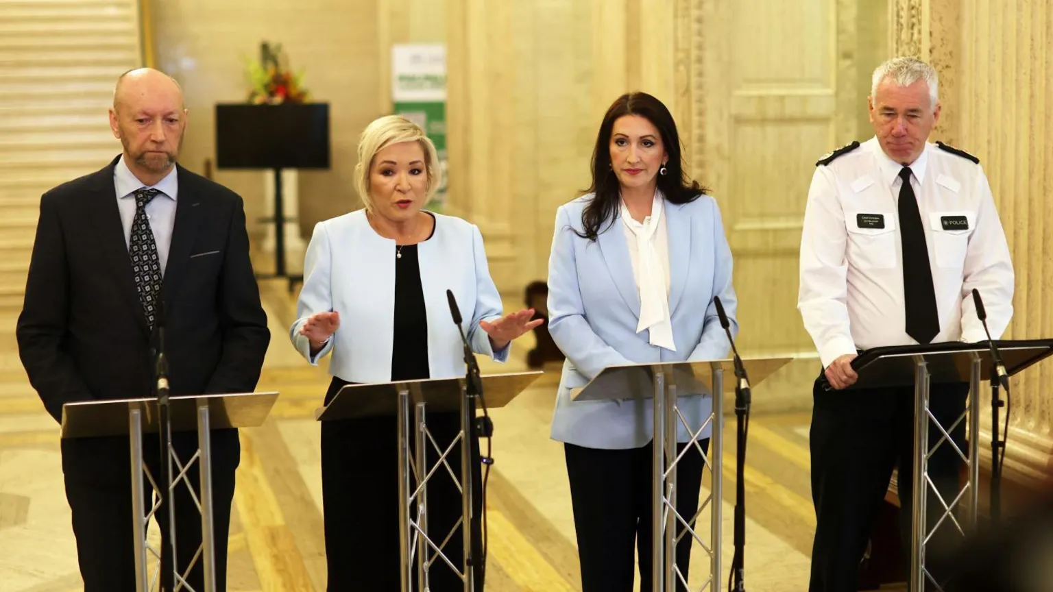  Four people stand behind lecterns with microphones, addressing a press briefing in a large, ornate indoor hall with marble walls and tall columns. All are dressed in formal or professional attire, and one person on the right wears a uniform with a visible police badge. 