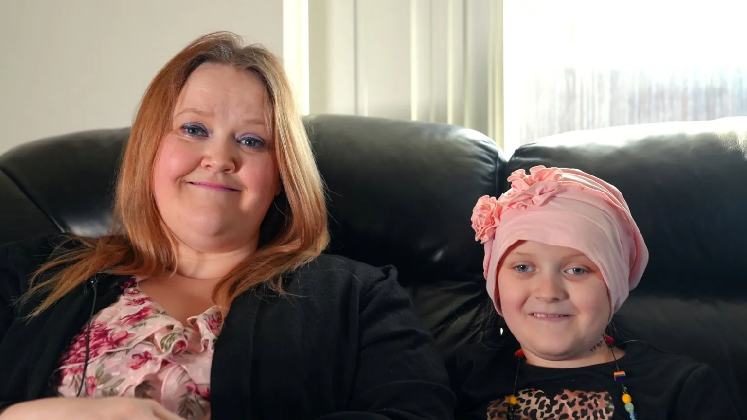 Sienna with her mum Emma. She has blue eyes and smiles at the camera. She wears a pink headwrap with flowers on it. Emma sits to the left of Sienna and smiles at the camera. She has blue eyes and ginger hair. 