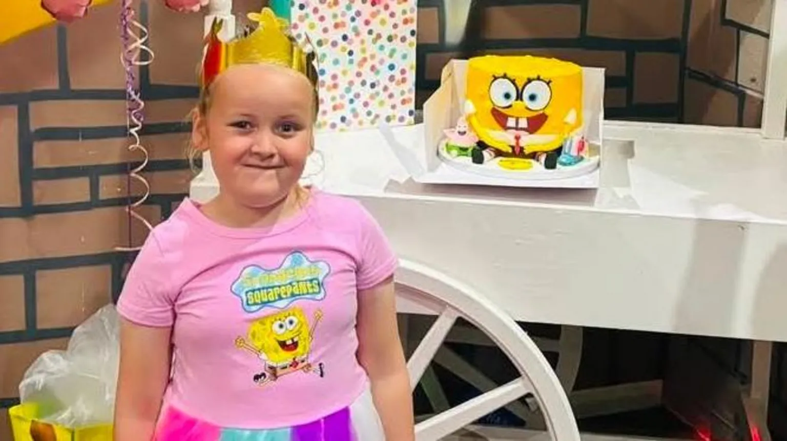 Jessica Gill Sienna pictured in a spongebob squarepants outfit at her birthday party. She wears a gold crown and stands in front of a spongebob square pants cake. 