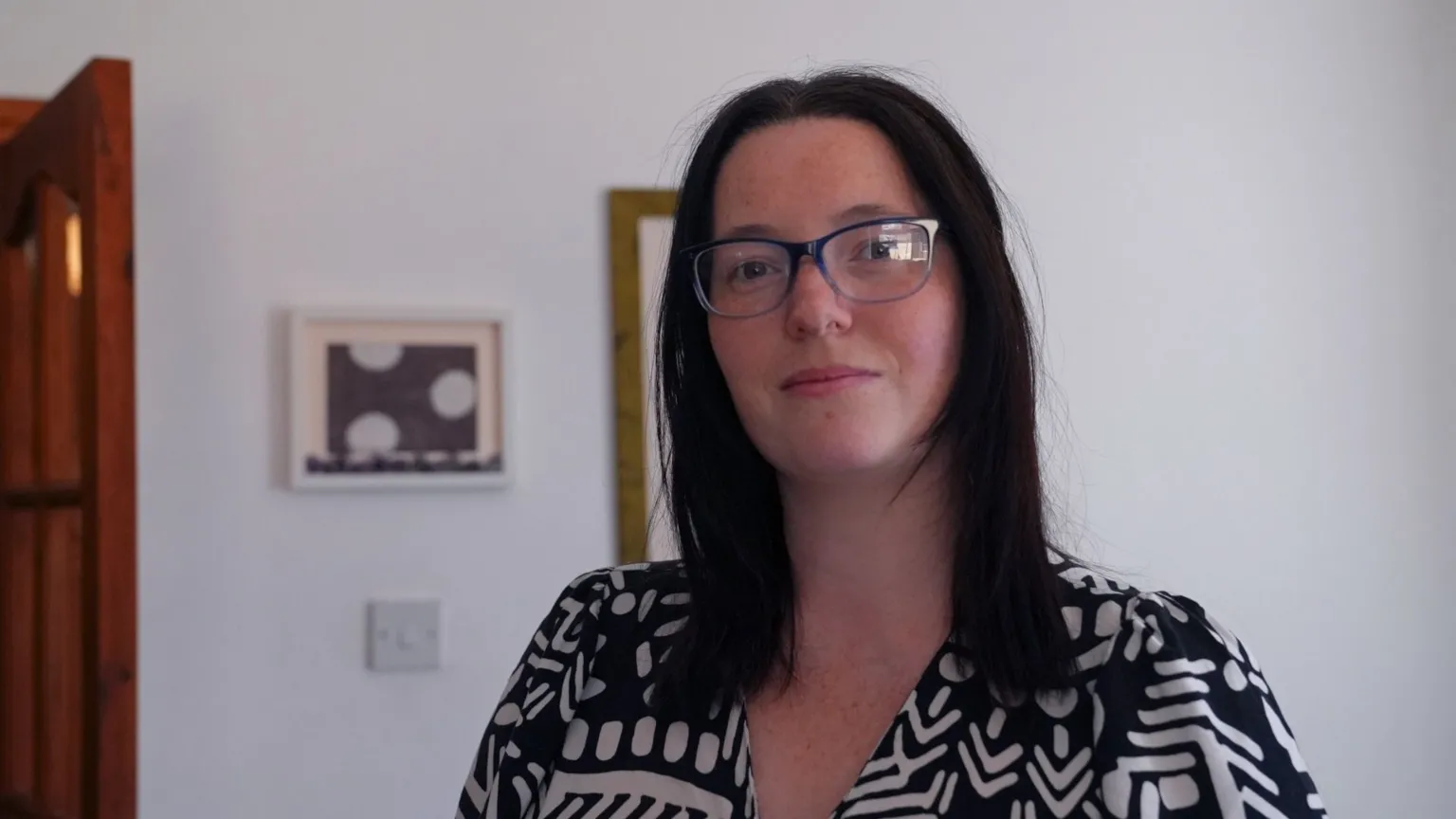 Jessica Gill stands infront of a white wall. She has black shoulder length hair, brown eyes and black square rimmed sunglasses. She looks directly at the camera. 