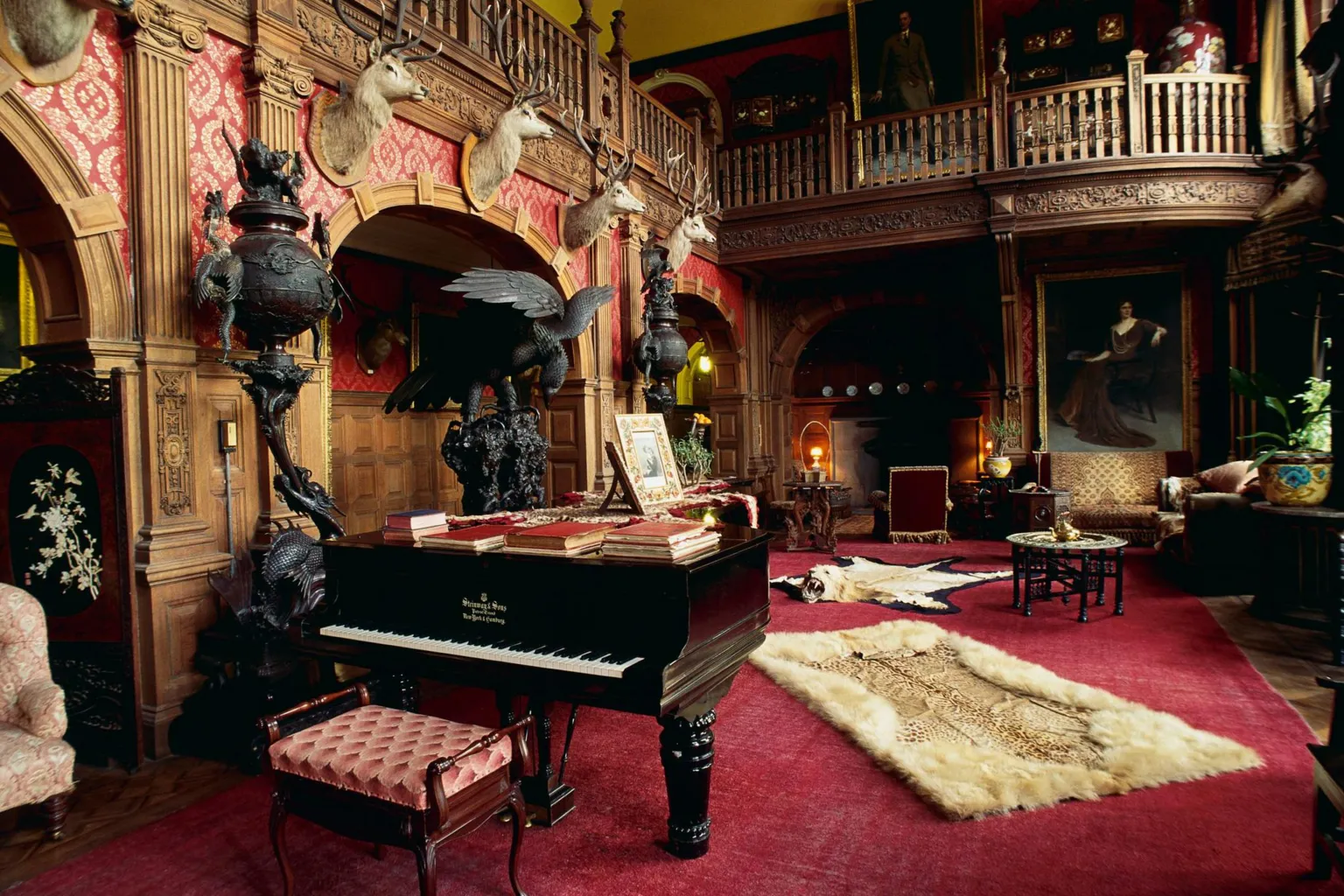  The image shows a large room inside the castle. There is a deep red carpet on the floor, a piano and stags' heads on a wall.