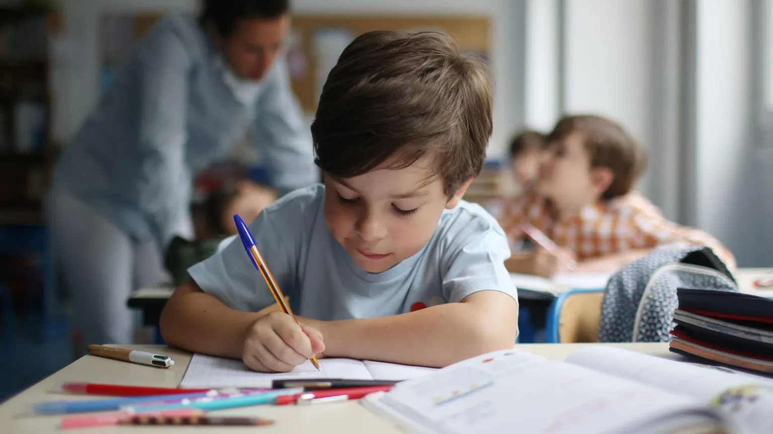  A child with short brown hair writing in a book at a desk in school.