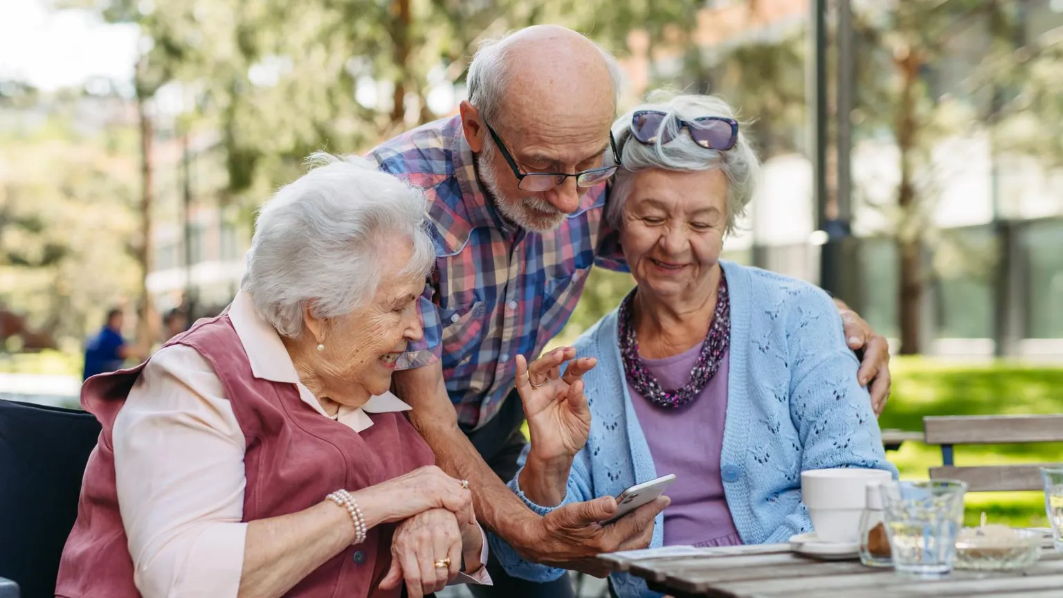  Three older people beside each other looking at a phone the man is holding. Both women have grey hair and the man is bald with short grey hair at the side.
