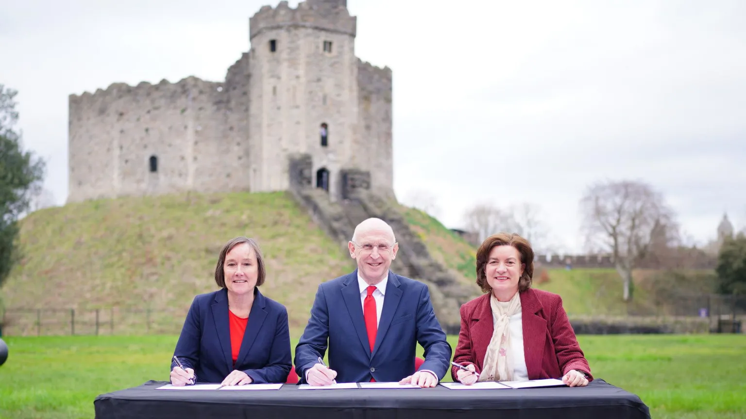  (i-r) Secretary of State for Wales Jo Stevens, Defence Secretary John Healey and First Minister of Wales Eluned Morgan during a visit to Cardiff Castle which can be seen in the background. They sit at a table with documents in front of them with pens poised to sign a defence deal.