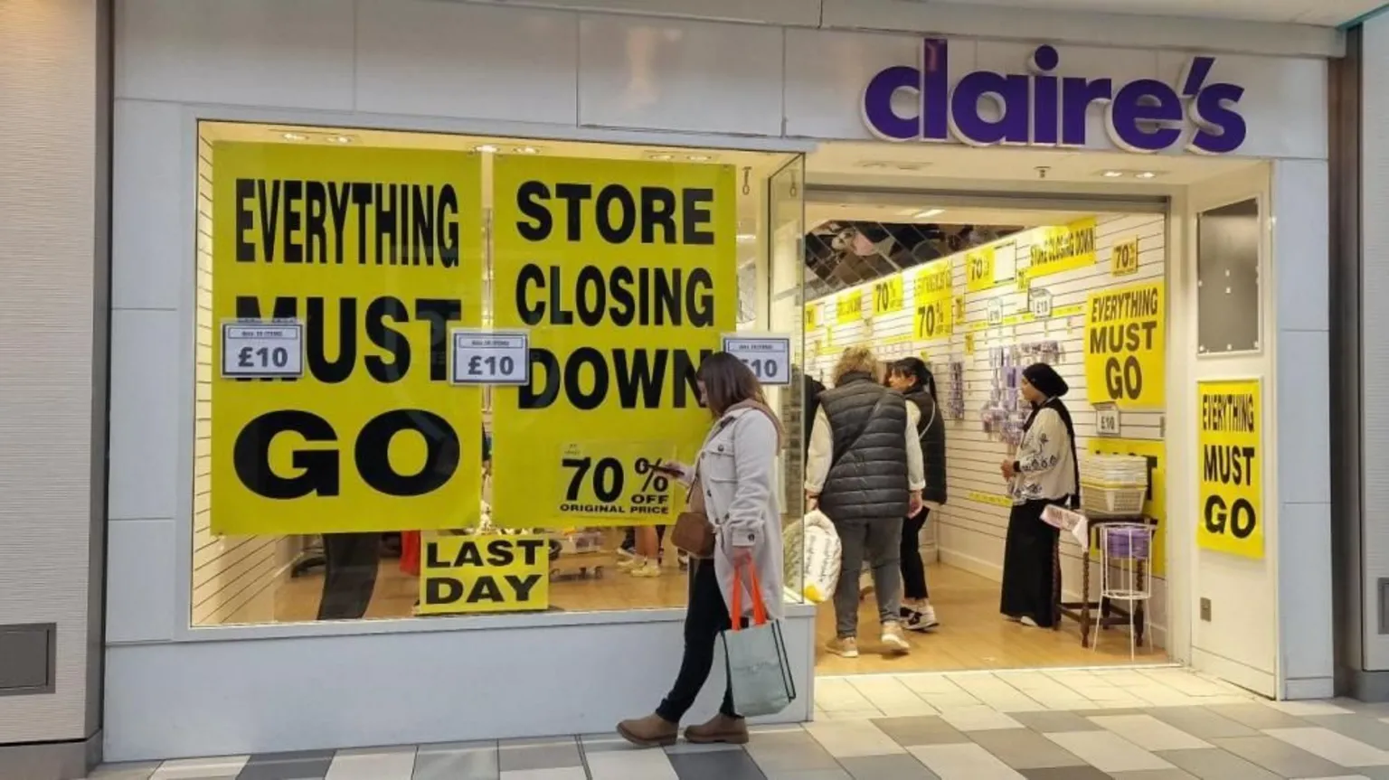 A woman looks at her phone outside a Claire's store, that has yellow and black closing down signs filling the windows saying 