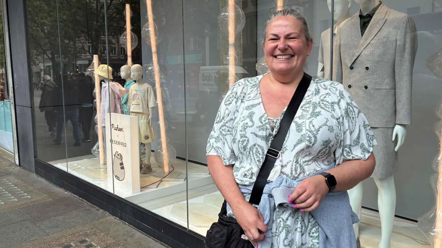 Silvia is smiling while standing in front of a shop window displaying mannequins. 