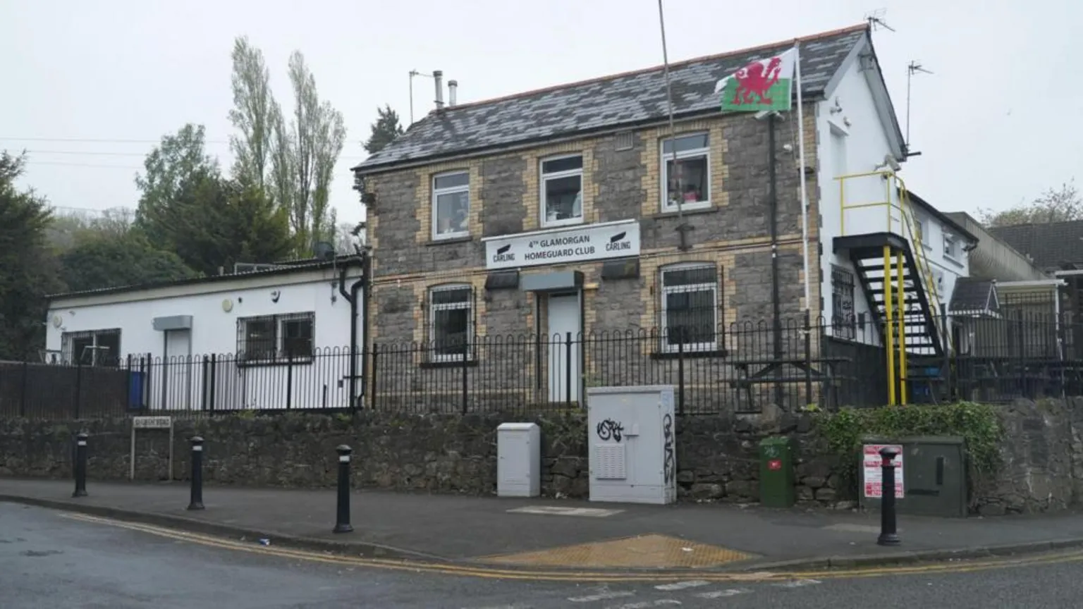 The Glamorgan Homeguard Club, a two storey grey-brown stone building, as seen from the street. There is a Welsh flag flying at full mast outside and black railings around it. A one-storey white building is to the left and there are stairs with yellow railings going up the right hand side of the club.